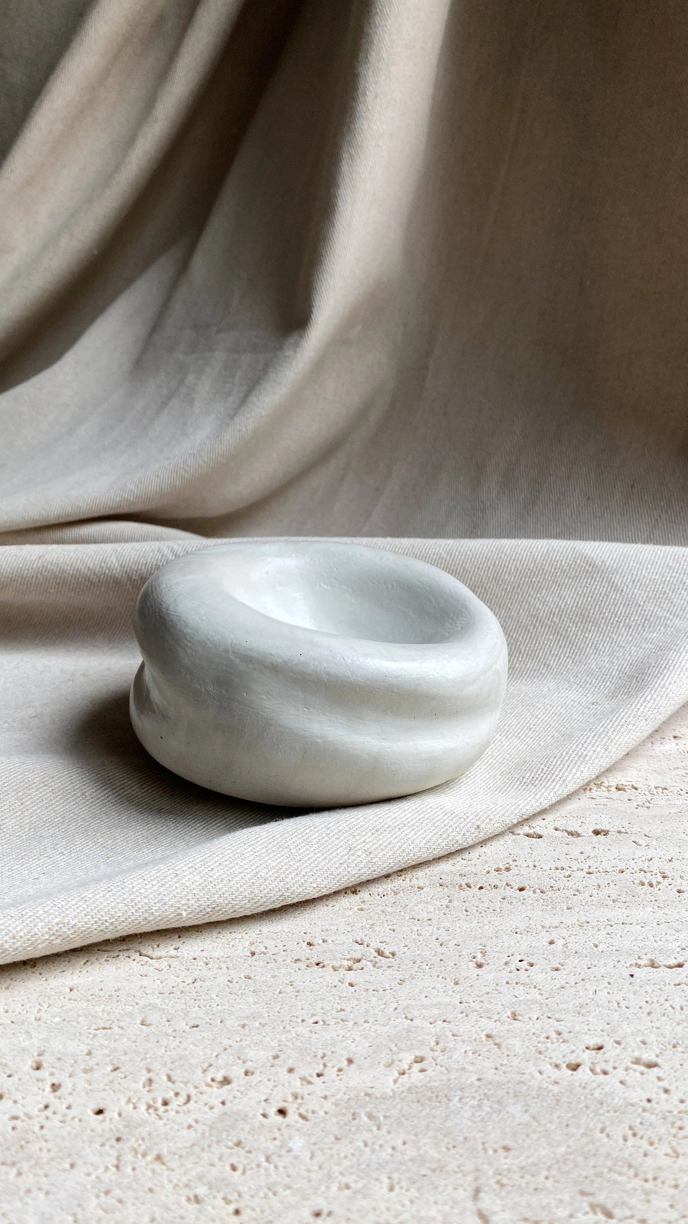 The Sumo Pocket Concrete Bowl by Faire, featuring a small, smooth, rounded and irregular design, rests on a textured stone surface in wabi sabi style with draped beige fabric in the background.