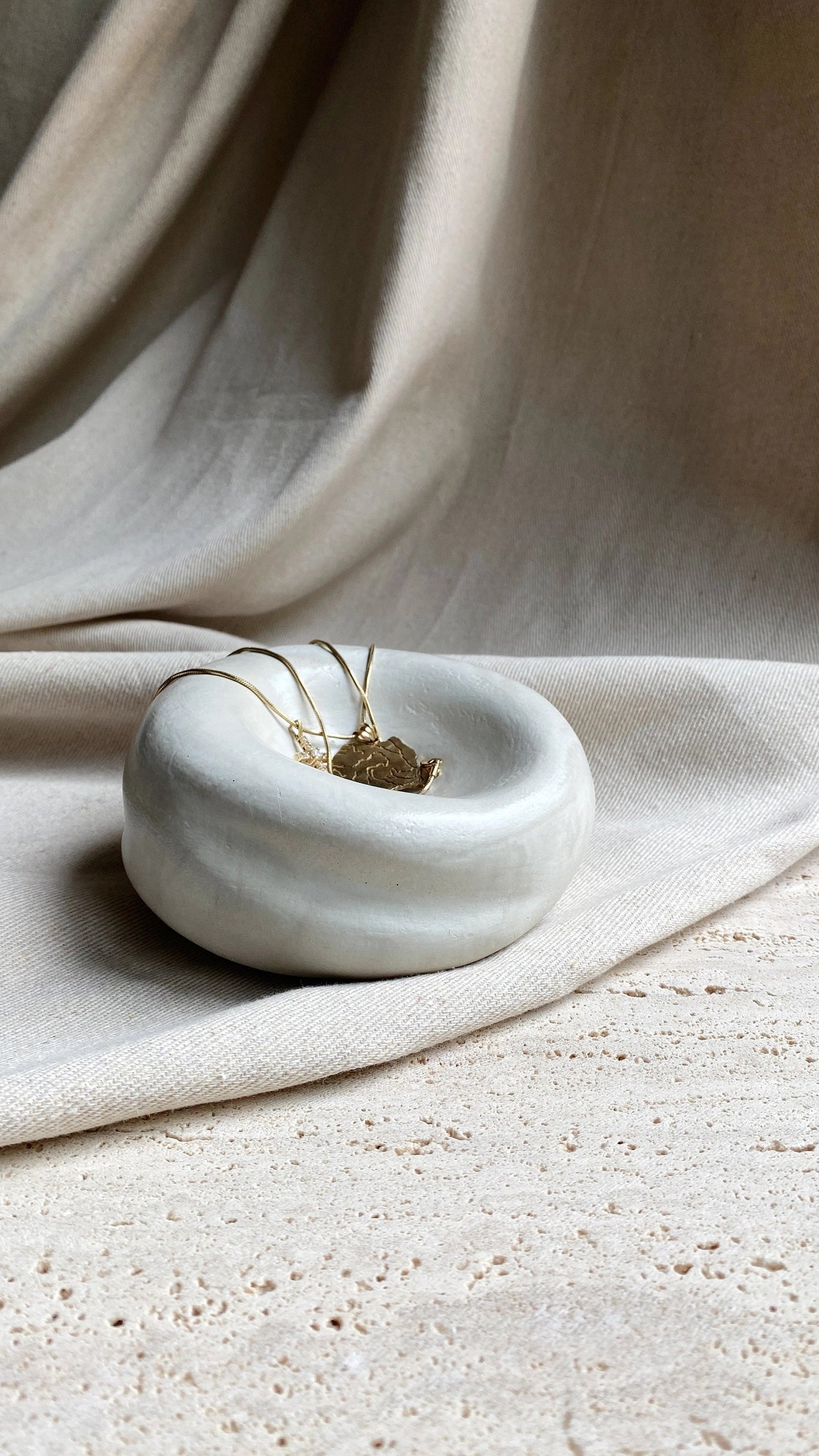 A pair of gold earrings sits in the smooth, round Sumo Pocket Concrete Bowl by Faire, placed on a textured stone surface with a beige fabric draped behind.