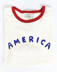 The Oat Collective America Puff Ringer Graphic T-Shirt features a white body, bold blue "AMERICA" puff print, red collar, and is crafted from premium Airlume cotton for comfort.