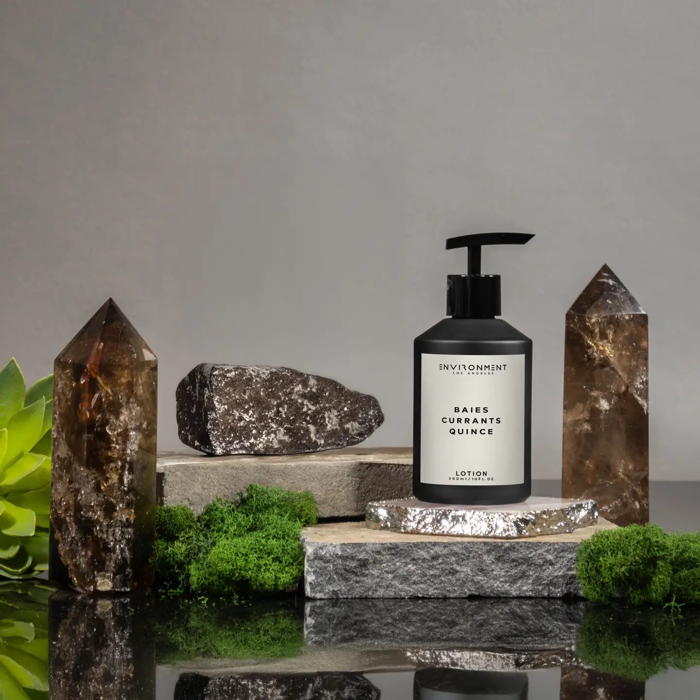 A black pump bottle of Faire Inspired Lotion sits on stone slabs with green moss, crystals, rocks, and a small plant, set against a neutral gray background.