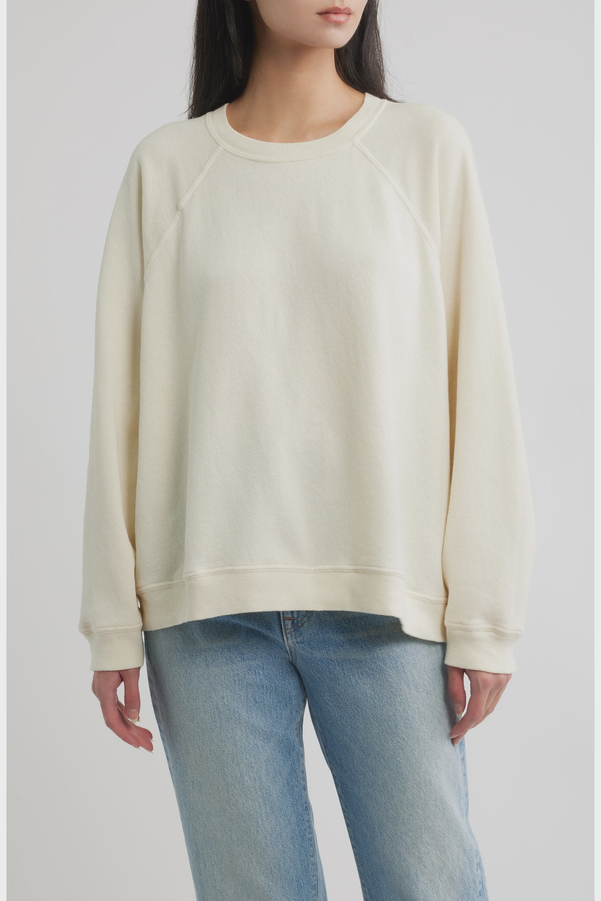 A person wearing The Rehearsal Sweatshirt in a loose, cream-colored raglan-sleeve style by The Great Inc., paired with light blue jeans, stands against a plain, light background. The person's face is partially out of frame.