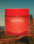 A red jar labeled "Magnesium Lavender Tallow Cream 4oz" by Faire is set against a desert with rocky formations and blue sky. The label features the brand name prominently, and the cream is infused with soothing lavender.