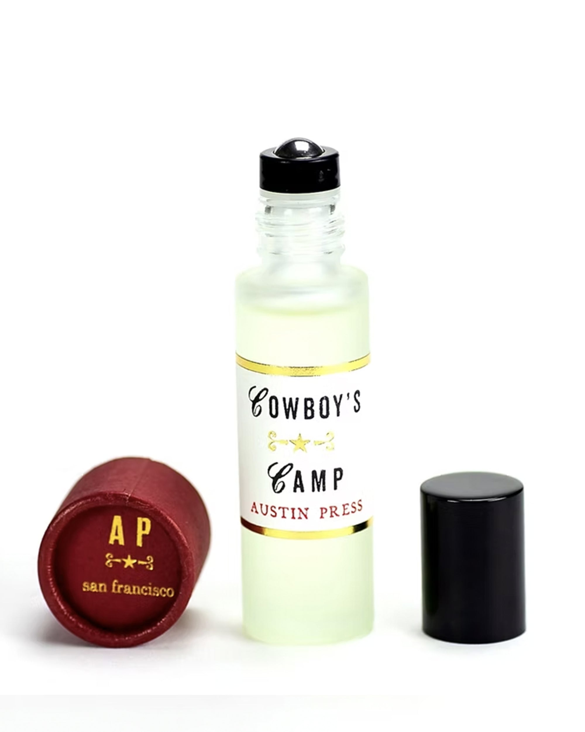 A small frosted glass bottle of Cowboy's Camp Perfume Oil by Faire stands upright, its black cap aside. A red cap with "AP san francisco" in gold lettering sits nearby, completing this unique fragrance’s presentation.