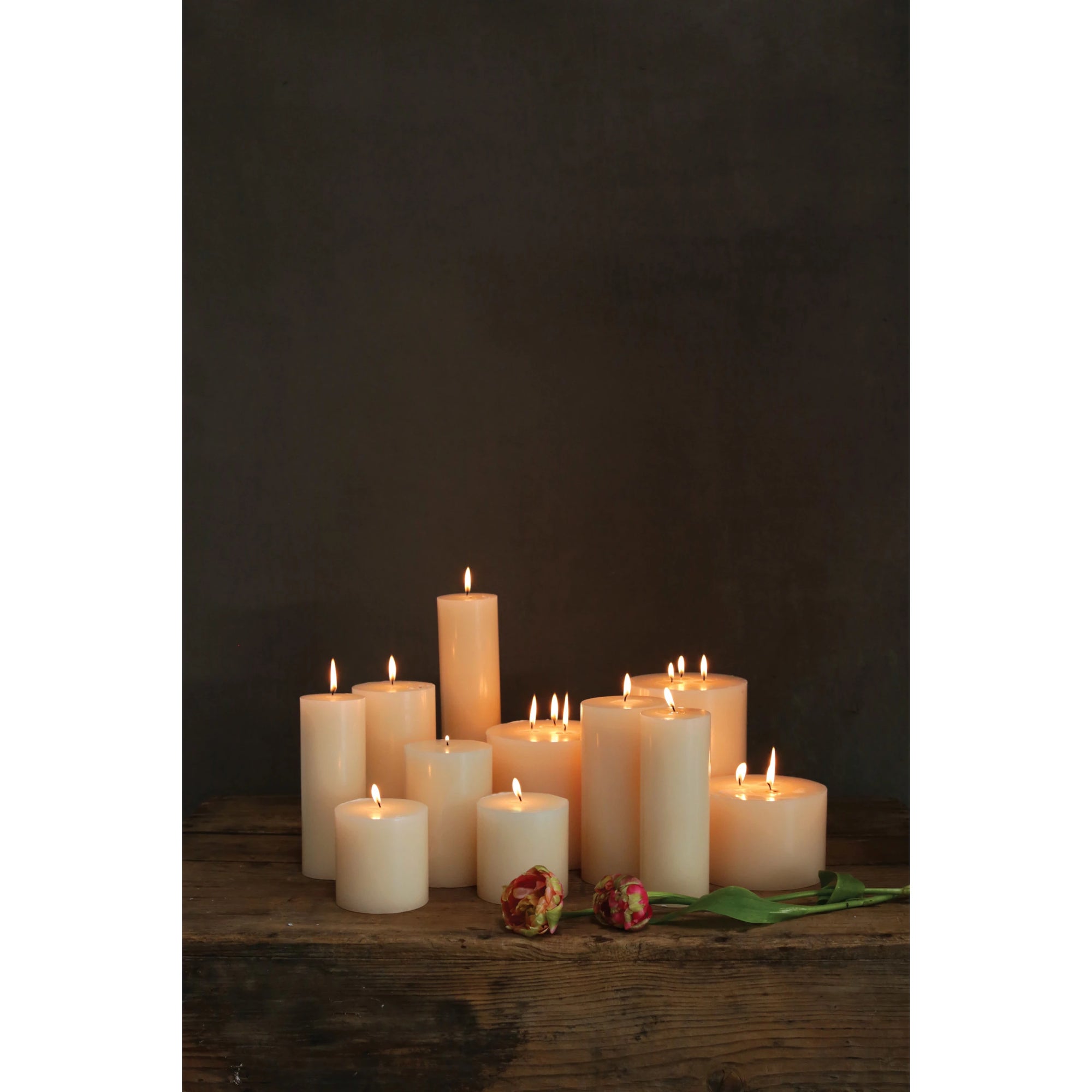 Creative Co-op's Unscented Candle 4x4, a white round pillar with up to 80 hours burn time, is arranged on a rustic wooden surface near two wilted red and yellow roses against a dark background.