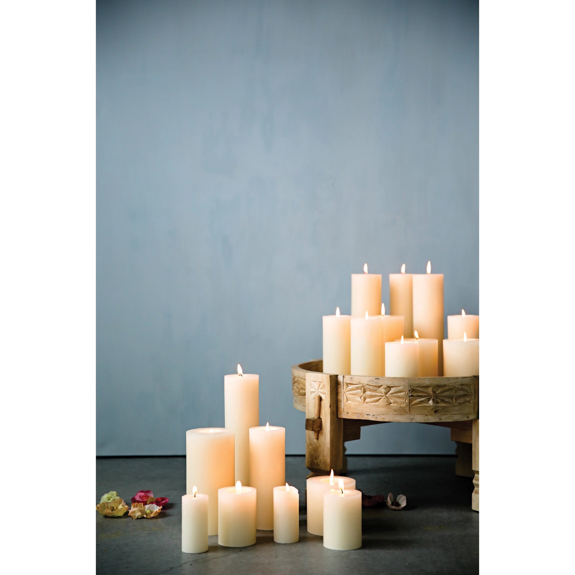 Creative Co-op Unscented Pillar Candles in varying heights, all lit, are arranged on the floor and a wooden pedestal with dried flowers nearby against a pale blue backdrop. Burn time: up to 145 hours.