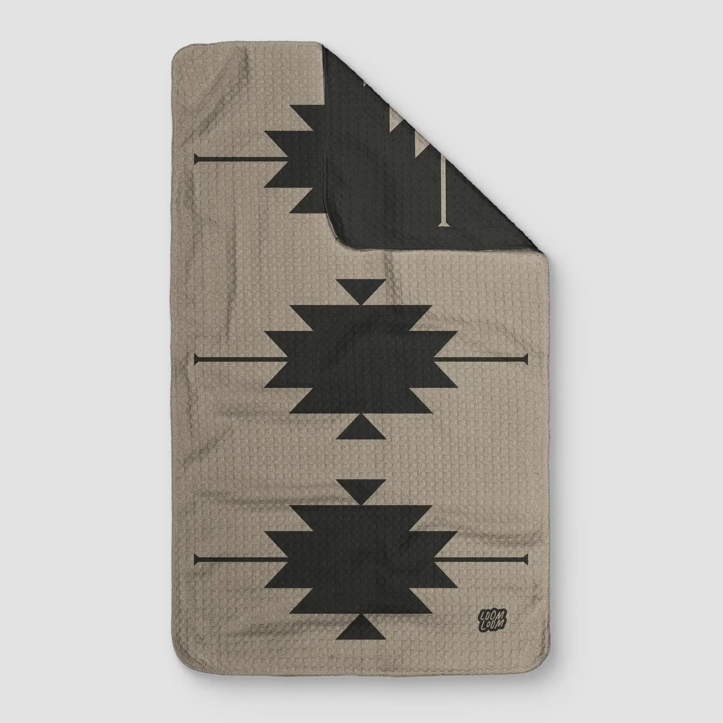 A gray rectangular SW Gray Towel by Faire, featuring a geometric black tribal pattern and a small "CAMP COZY" logo in the bottom right corner, is shown with the top right corner folded to reveal the reverse side.