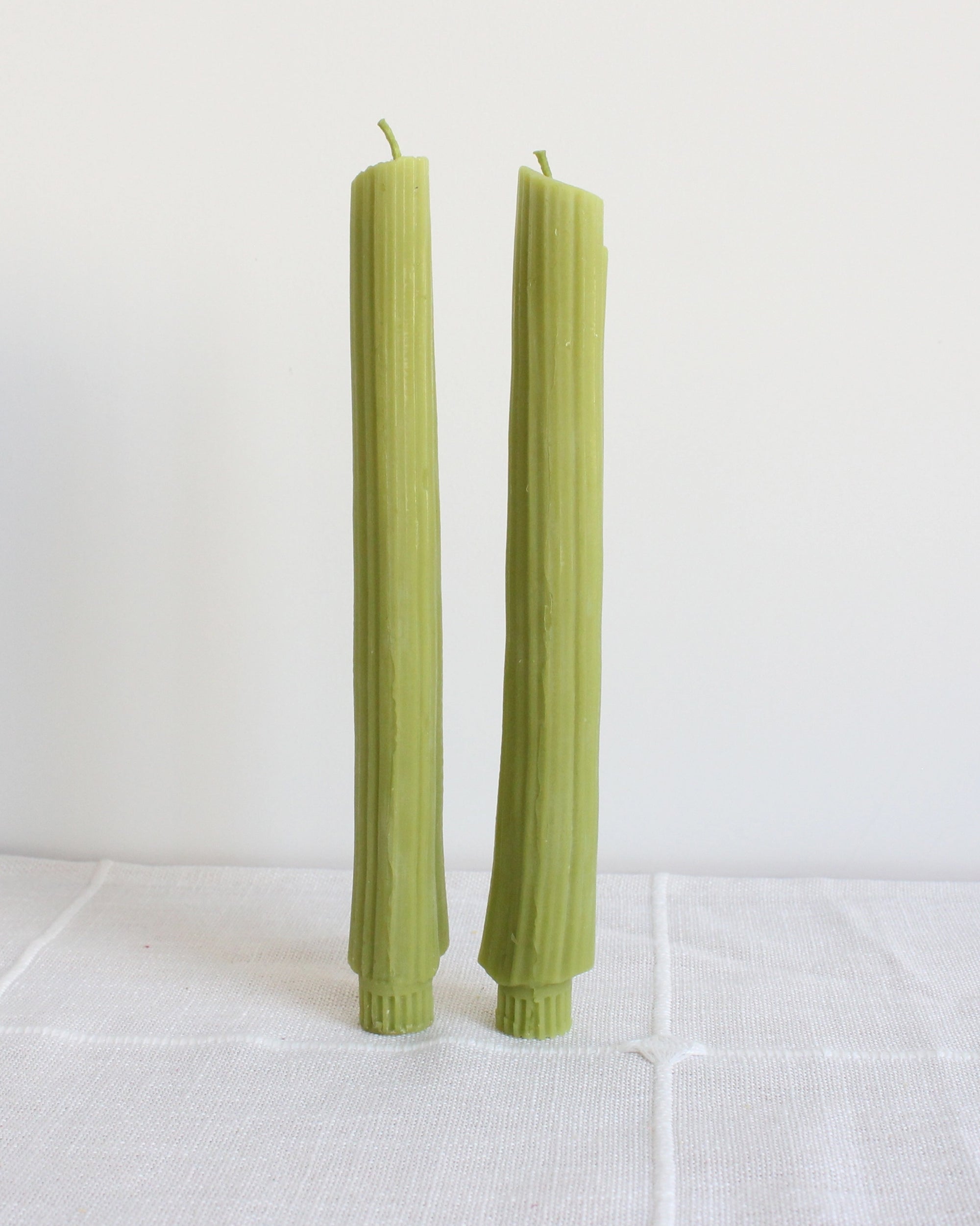 Two Faire Celery Beeswax Candles, handmade in Los Angeles, stand upright on a white cloth against a plain white background.