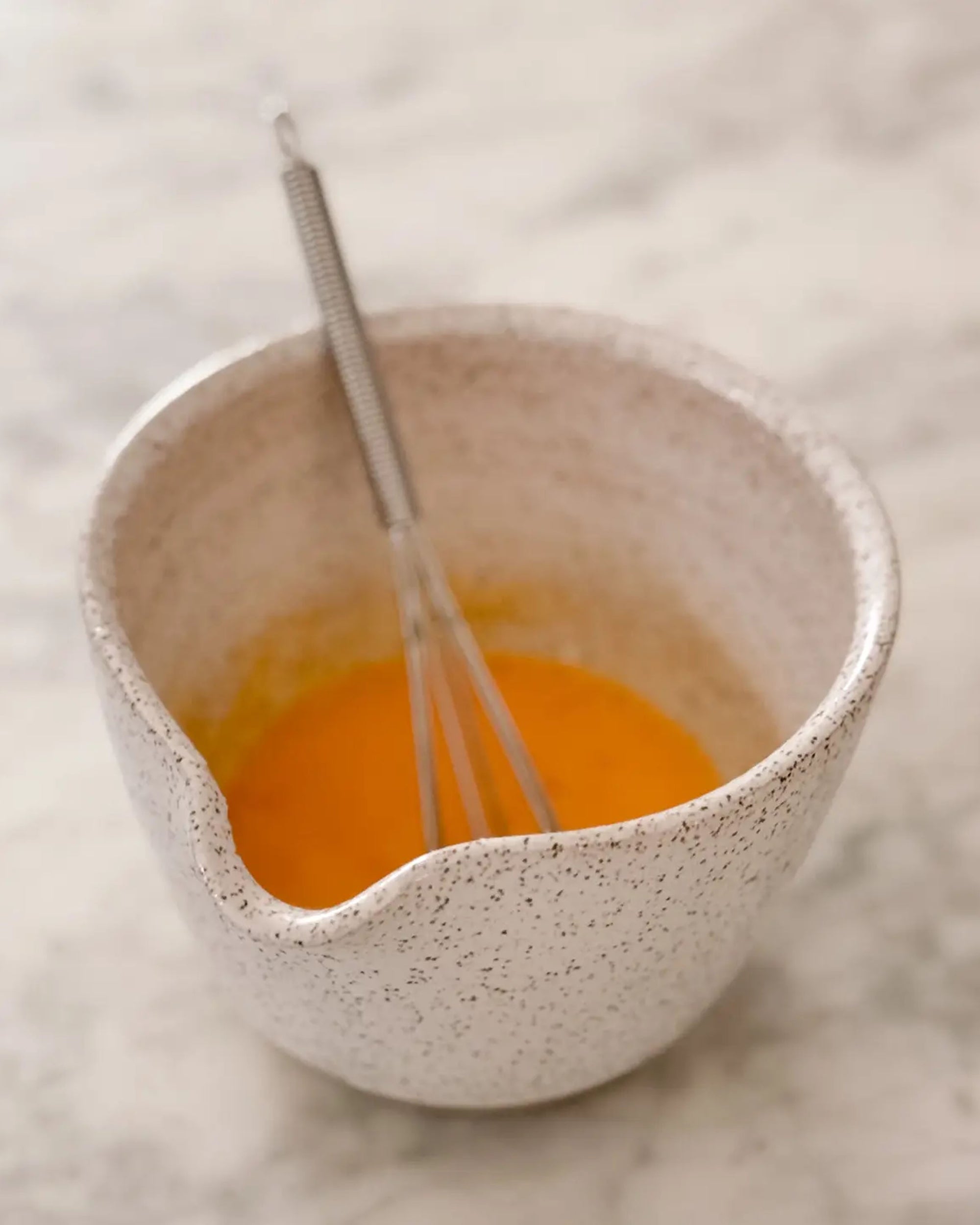 The Faire Ceramic Bowl w/ Whisk, featuring a handmade speckled design and pour spout, holds a yellow-orange liquid being mixed on a light marble surface.