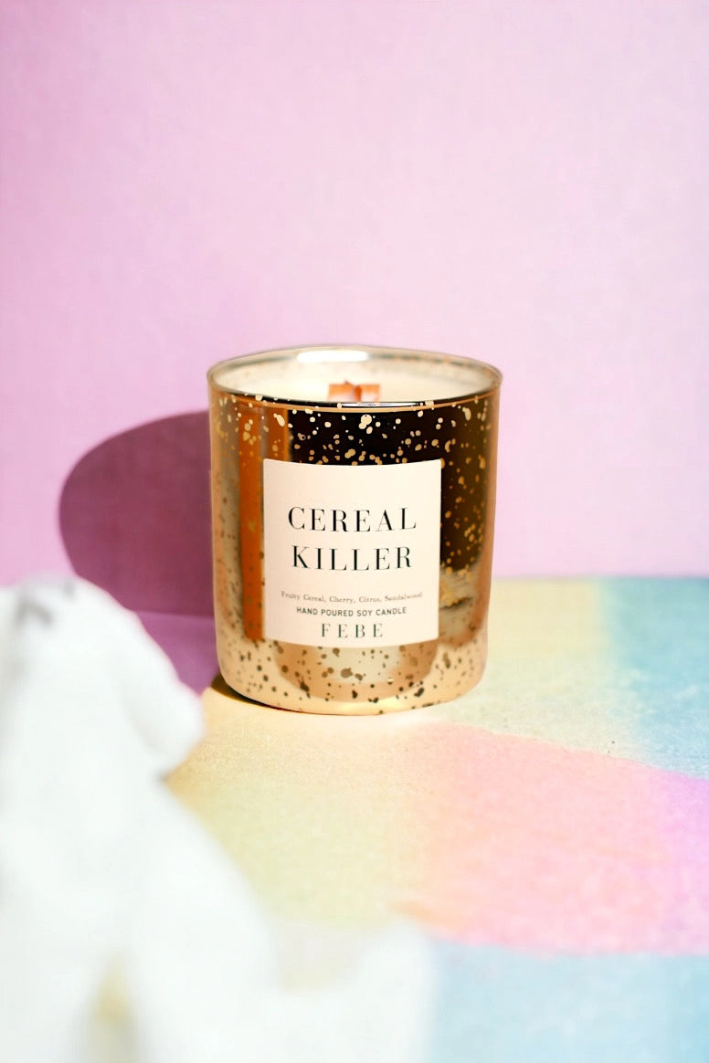 A FEBE candle in a speckled container labeled &quot;CEREAL KILLER&quot; stands on a surface with pink and yellow hues in the background, casting colorful shadows in Arizona style.