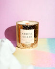 A FEBE candle in a speckled container labeled "CEREAL KILLER" stands on a surface with pink and yellow hues in the background, casting colorful shadows in Arizona style.