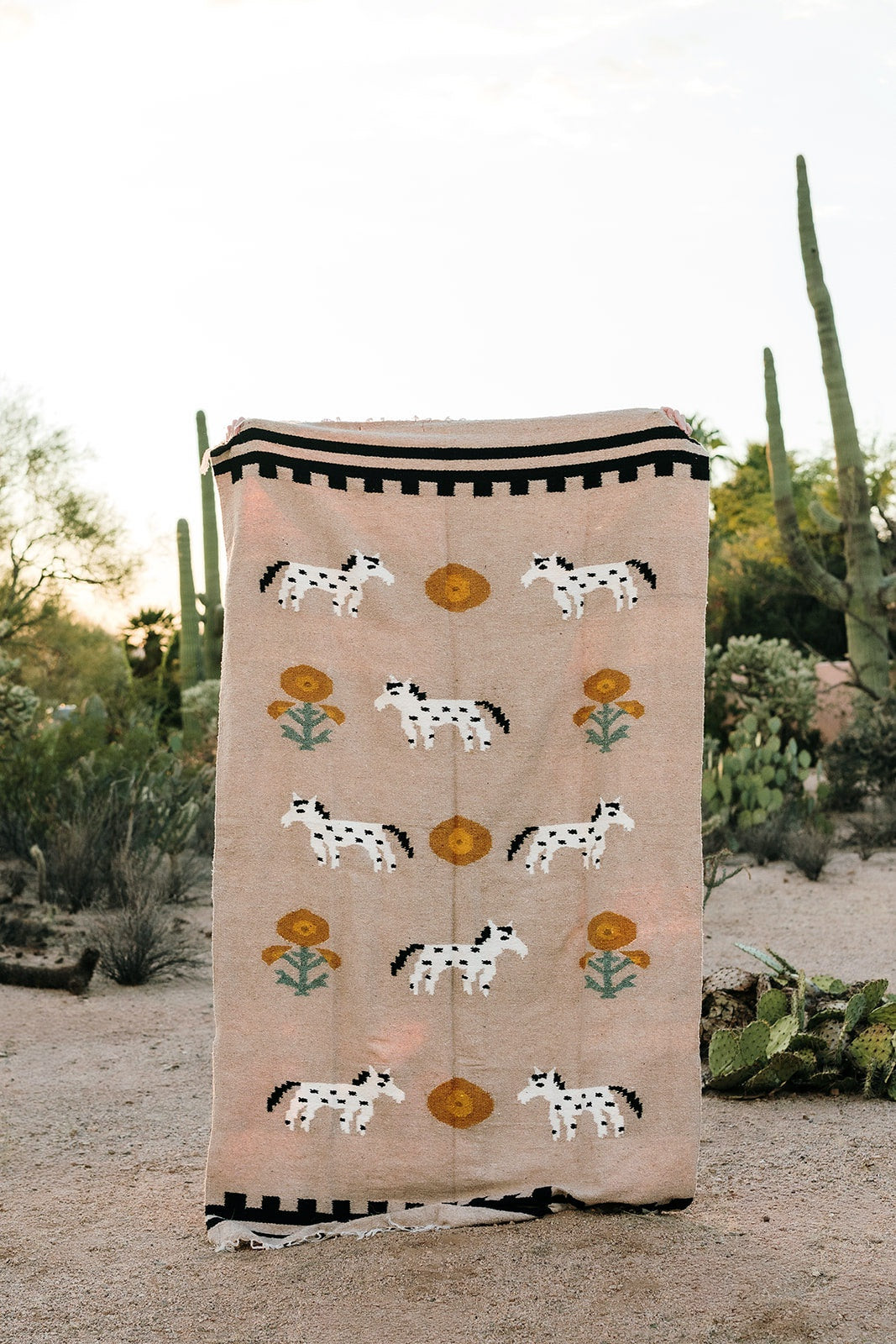 The TT Handwoven Blanket Poppies n Ponies by Faire, featuring white horses with black spots and yellow flowers, is held upright in a desert landscape with sand, cacti, and shrubs in the background.