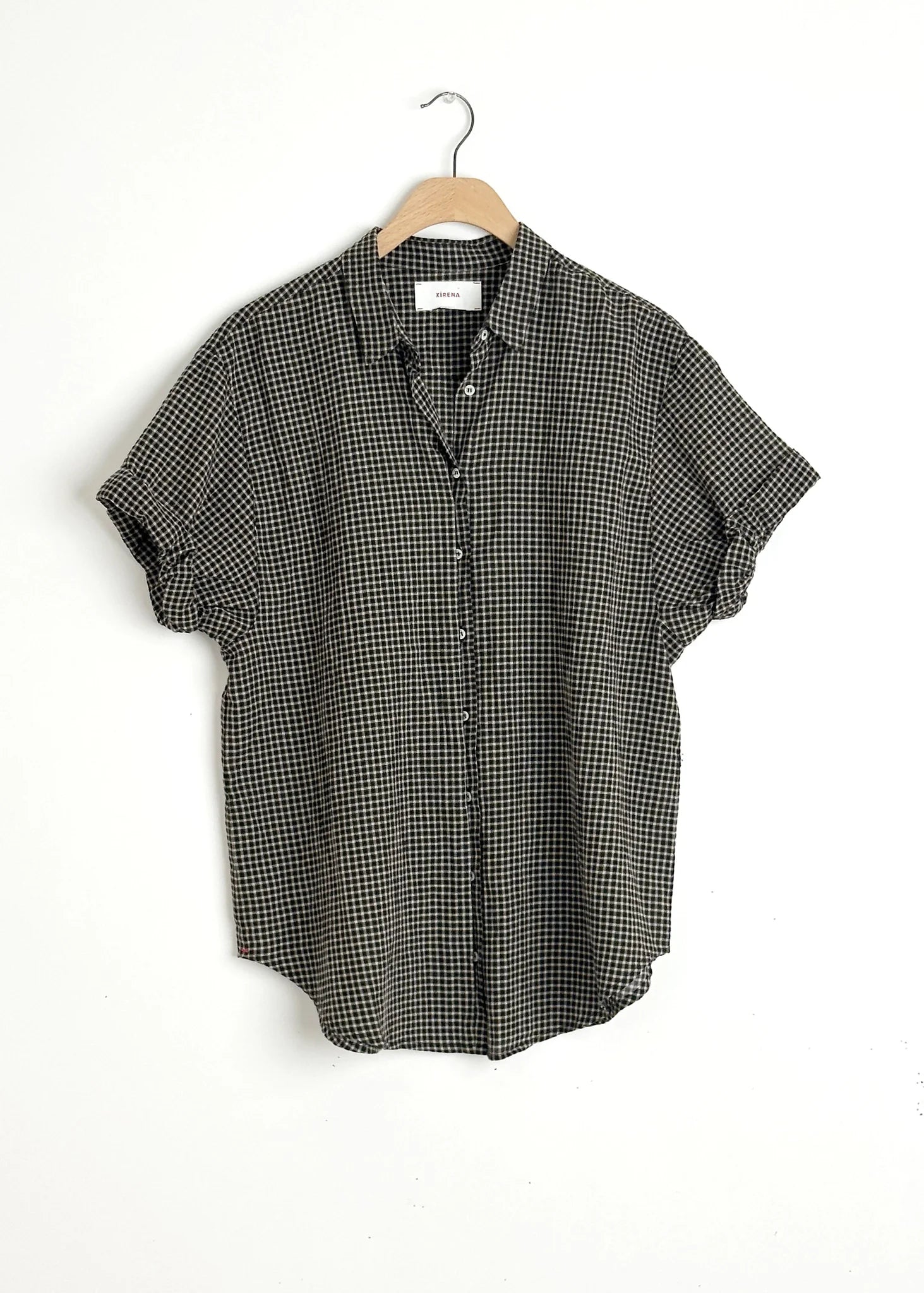 The Xirena Channing Shirt S25, a short-sleeved black and white checkered cotton chambray piece, hangs on a wooden hanger against a plain white wall.