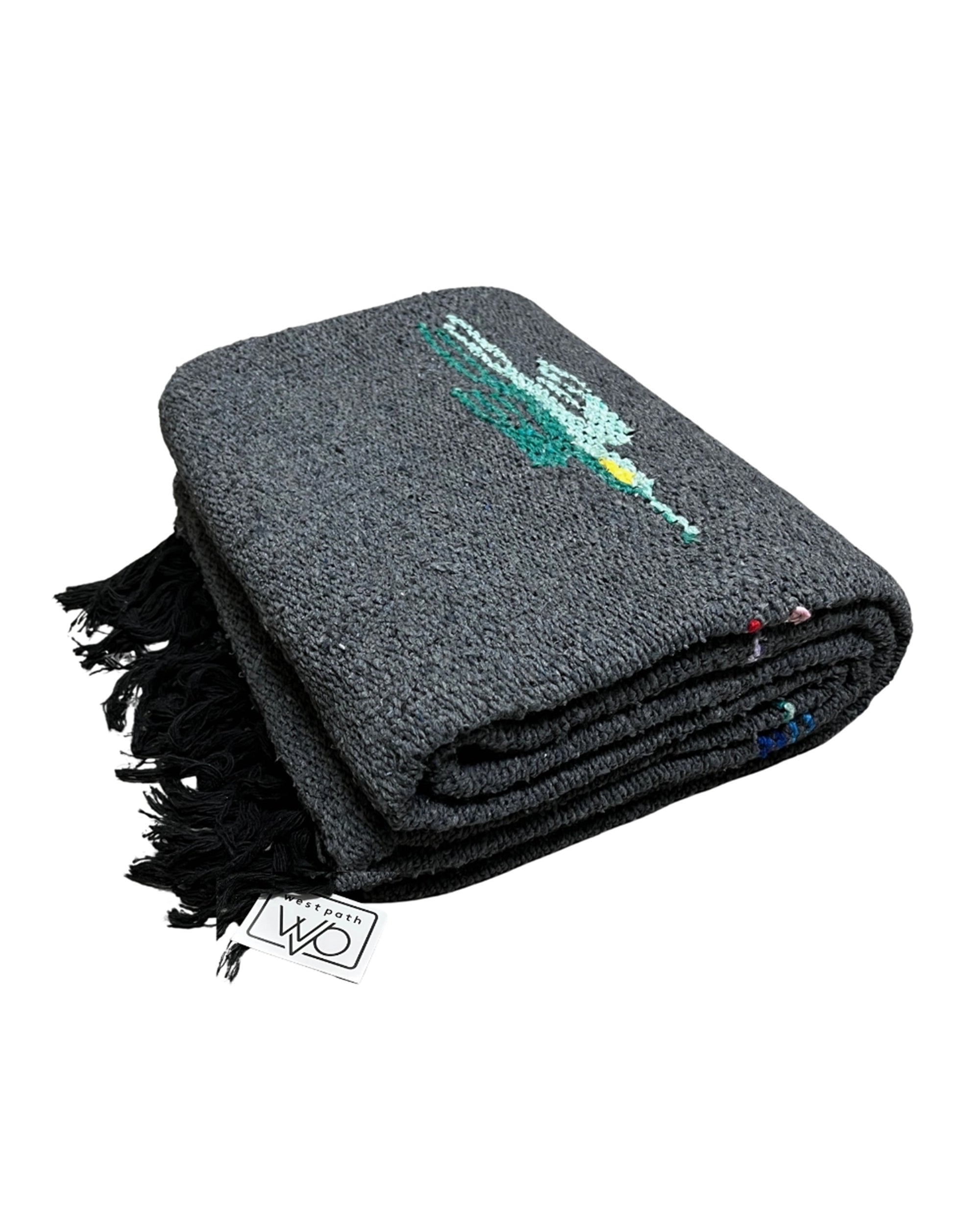 A folded Charcoal Thunderbird Baja Blanket by Faire features a dark gray woven design with black fringe, subtle colorful patterns on top, and a visible branded tag highlighting its handmade quality in Mexico.