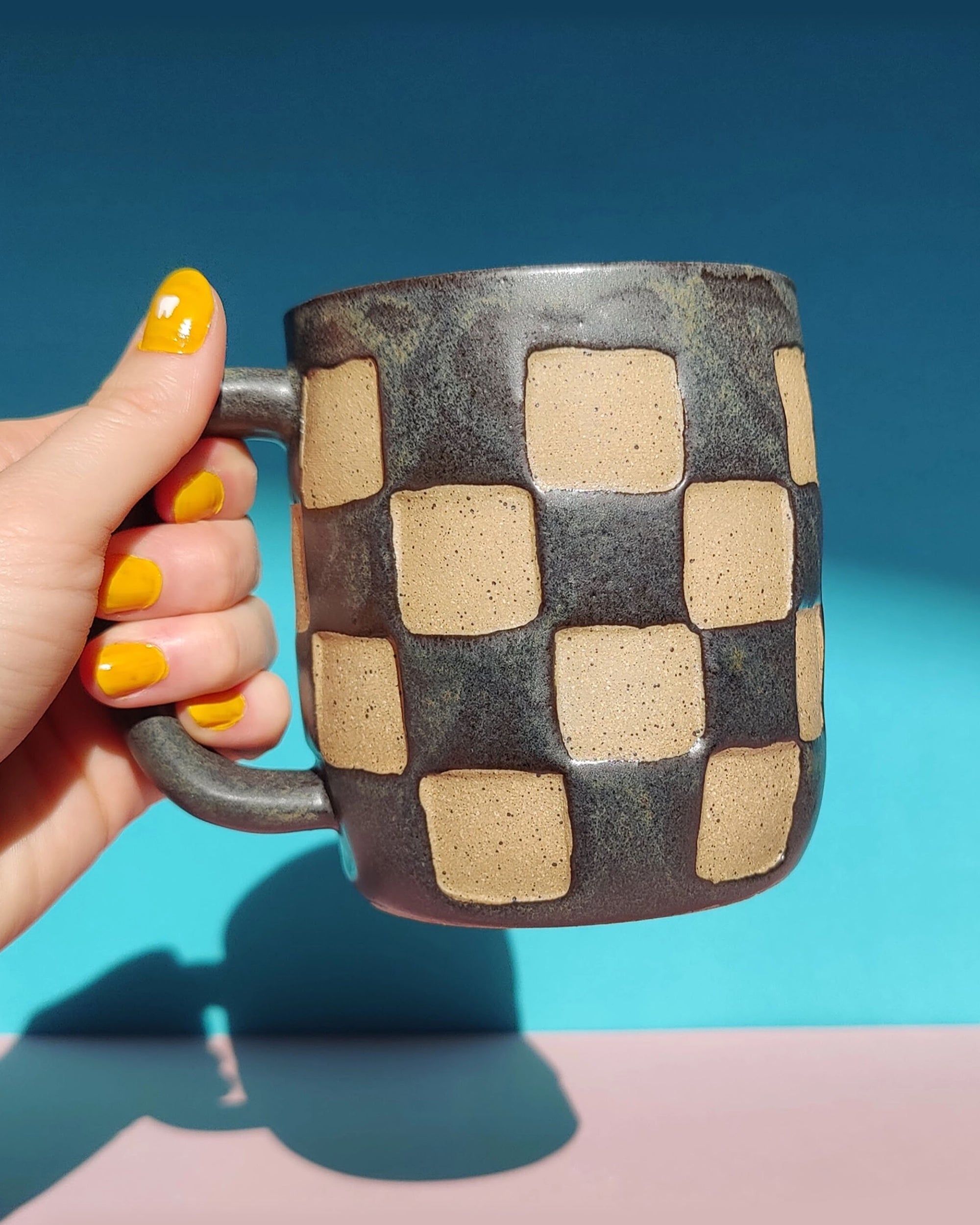 A hand with bright yellow nail polish holds the OC Checkerboard Mug by Faire, which features a beige and dark gray pattern, set against a blue and pink background.