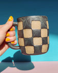 A hand with bright yellow nail polish holds the OC Checkerboard Mug by Faire, which features a beige and dark gray pattern, set against a blue and pink background.