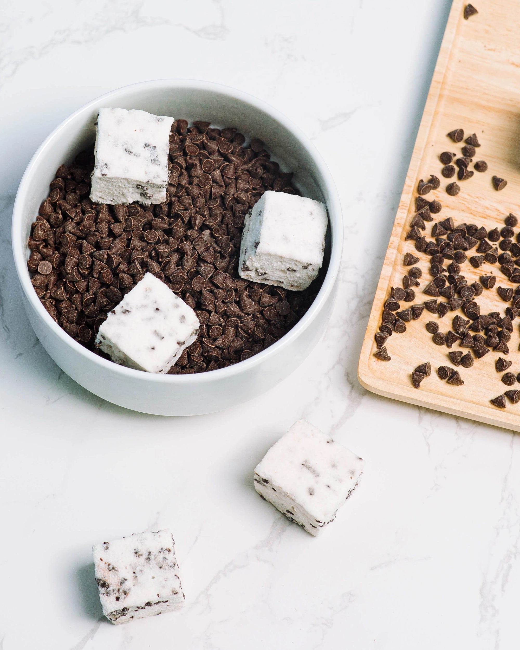 A white bowl holds Faire Choc Chip Marshmallows topped with more chocolate chips. Nearby, a wooden board has extra chocolate chips and a dairy-free Choc Chip Marshmallow from Faire rests on the table.