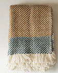 The Faire Chunky Twill Throw, with brown and blue diagonal stripes and a thick cream fringe along the edge, is folded and displayed on a white surface.