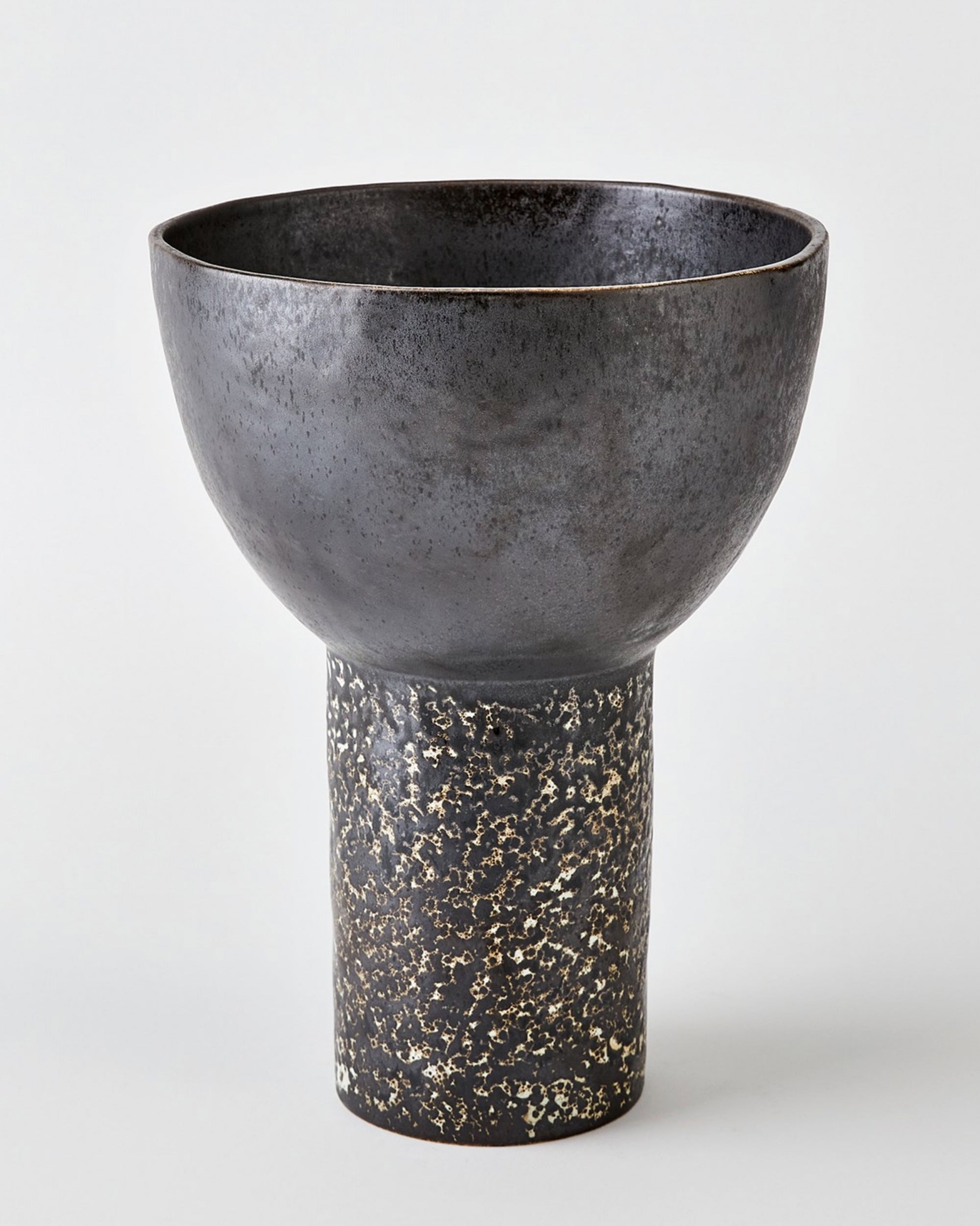 The 7.10712 Cinder Footed Bowl LG by Global Views features a wide, dark matte bowl atop a textured, speckled cylindrical base crafted from Portuguese stoneware and displayed against a plain light background.