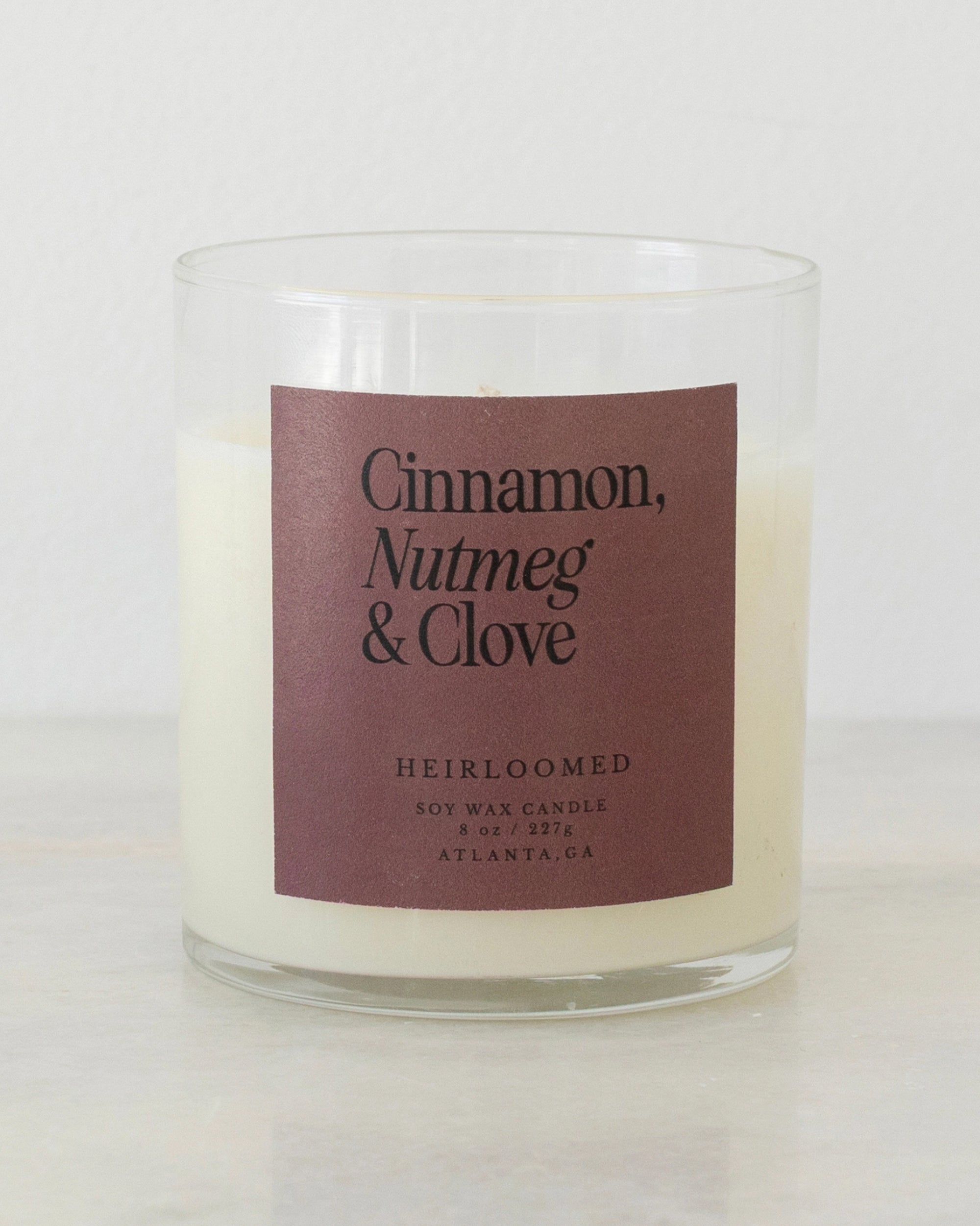 A Heirloomed Candle by Faire sits on a light surface, featuring a glass jar, brown label with “Cinnamon, Nutmeg & Clove,” and product details: Soy Wax Candle 8oz 227g Atlanta, GA, set against a plain background.