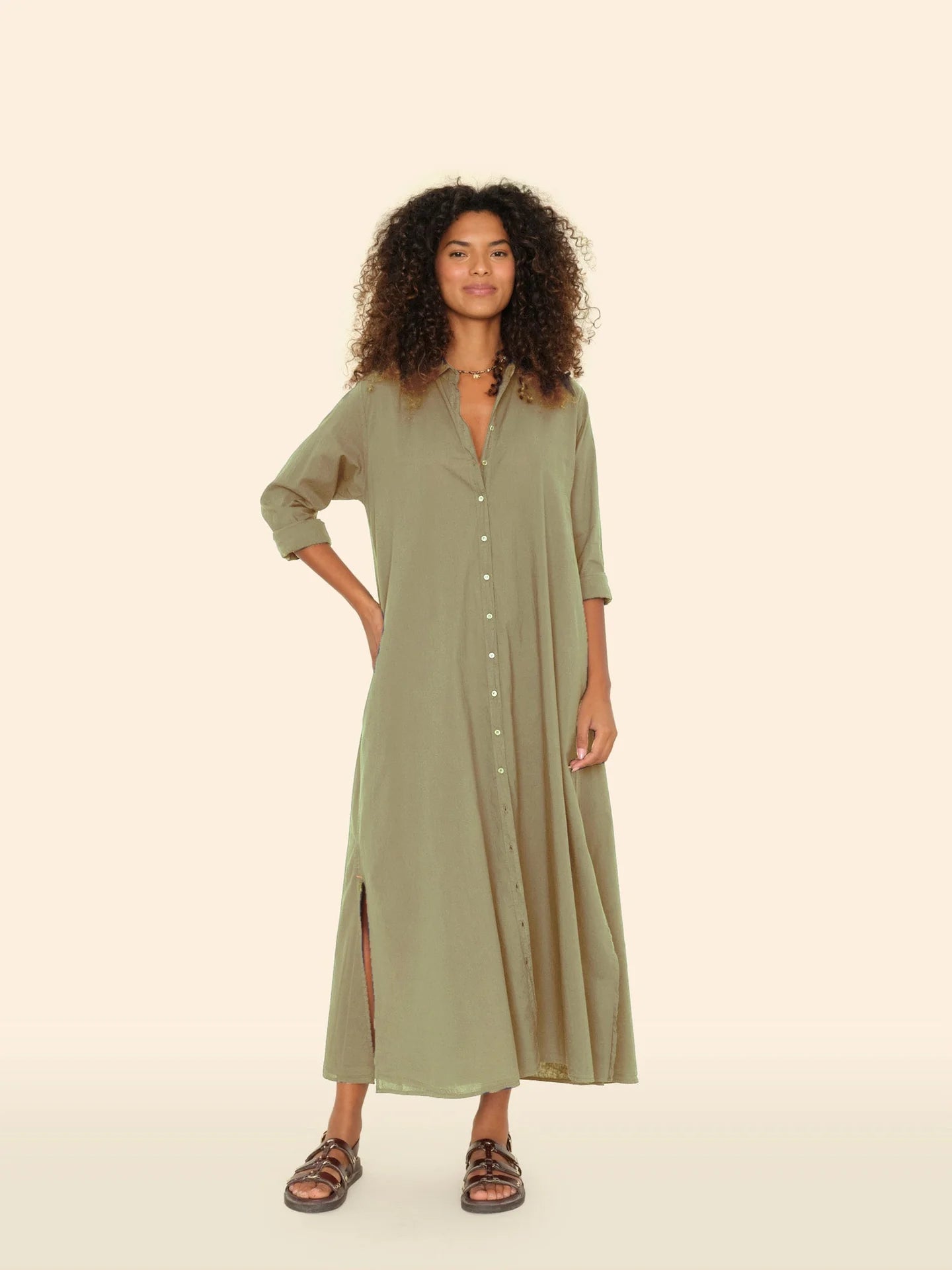 With confidence, a curly-haired person wears the Xirena Boden Dress, an olive-green cotton poplin shirtdress with side slits. They pair it with dark sandals and a necklace, posing against a beige backdrop.