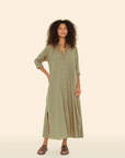 With confidence, a curly-haired person wears the Xirena Boden Dress, an olive-green cotton poplin shirtdress with side slits. They pair it with dark sandals and a necklace, posing against a beige backdrop.