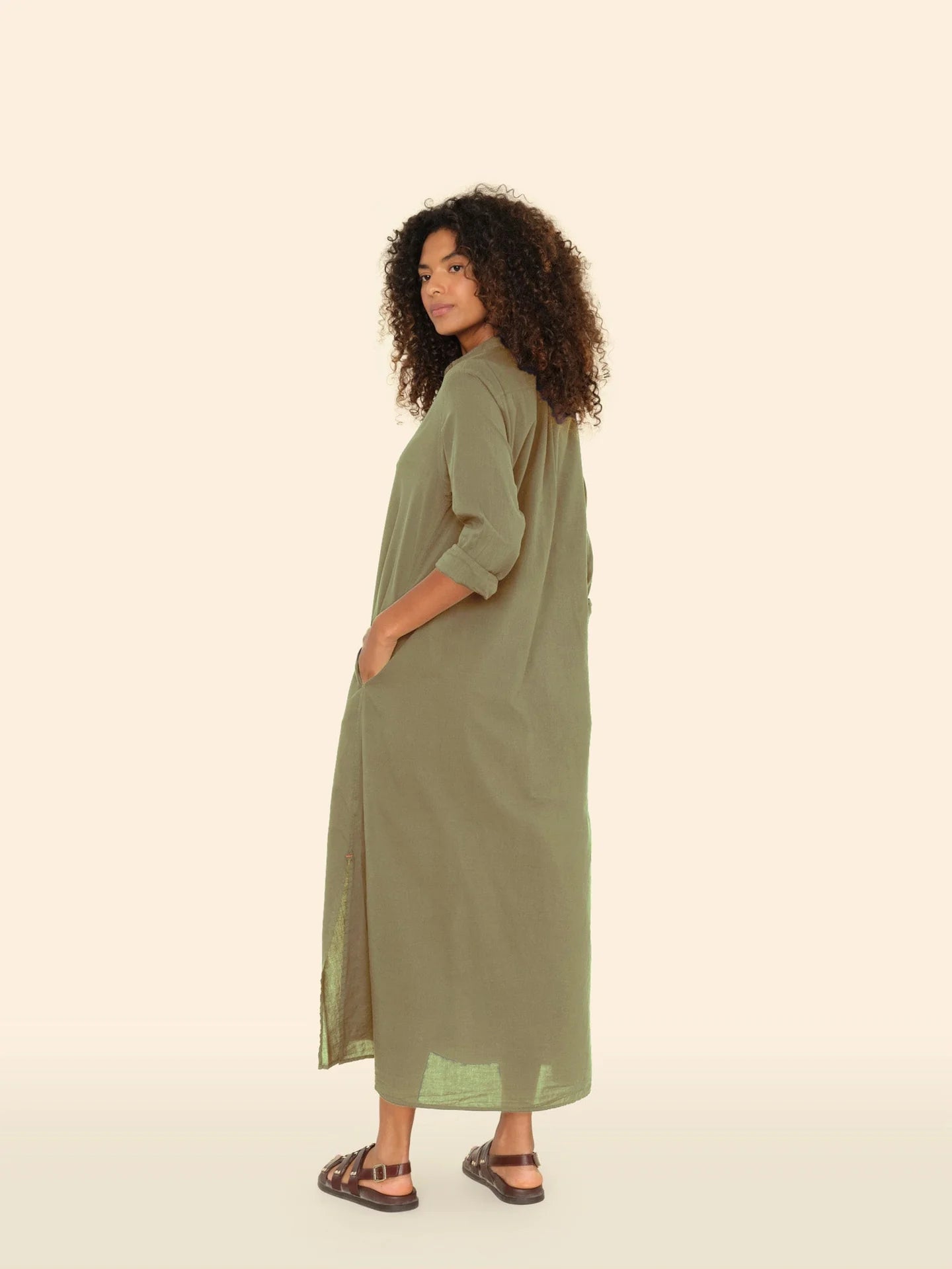 With curly hair, a person stands sideways and glances over their shoulder. They wear Xirena's Boden Dress, an olive green shirtdress with rolled-up sleeves. Paired with sandals, the cotton poplin fabric adds casual elegance against a light, neutral background.