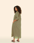 With curly hair, a person stands sideways and glances over their shoulder. They wear Xirena's Boden Dress, an olive green shirtdress with rolled-up sleeves. Paired with sandals, the cotton poplin fabric adds casual elegance against a light, neutral background.