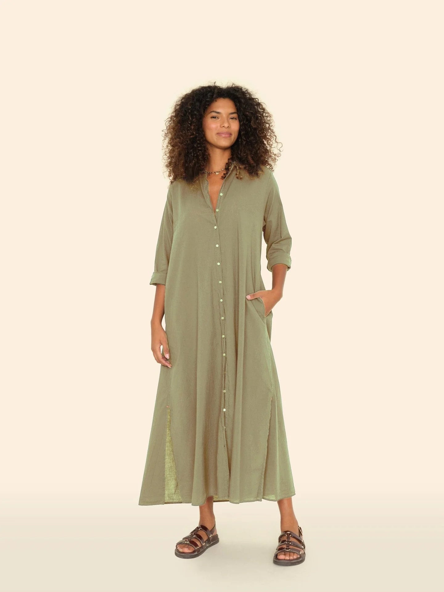 A person with curly hair is wearing a Xirena Boden Dress, a tailored light olive green shirtdress made from cotton poplin. Their elbows showcase rolled-up sleeves as they stand against a neutral backdrop, donning brown sandals and smiling, with one hand casually in their pocket.