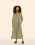 A person with curly hair is wearing a Xirena Boden Dress, a tailored light olive green shirtdress made from cotton poplin. Their elbows showcase rolled-up sleeves as they stand against a neutral backdrop, donning brown sandals and smiling, with one hand casually in their pocket.