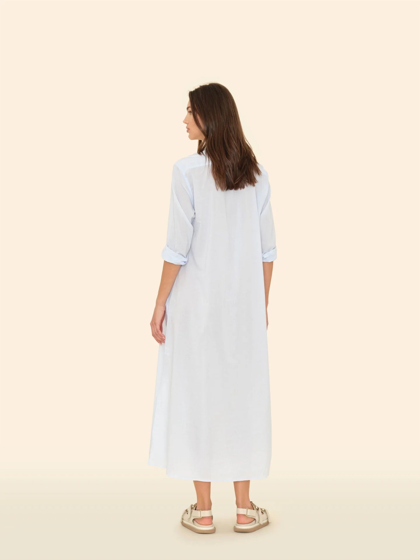 A person with long hair faces away, wearing the Boden Dress by Xirena in light blue cotton poplin with rolled-up sleeves. They pair it effortlessly with beige sandals against a solid, light beige background.