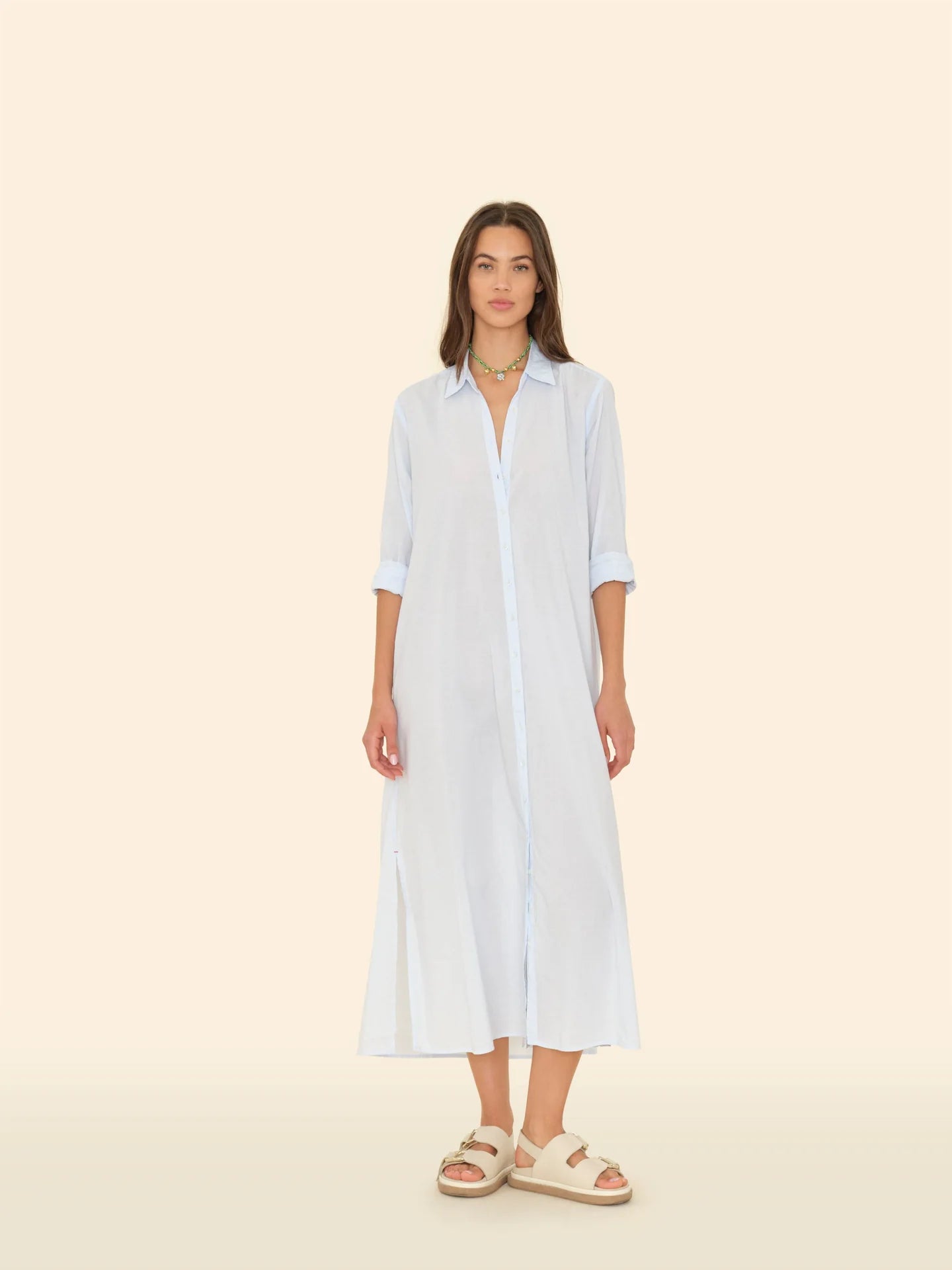 A person in a light blue Boden Dress from the brand Xirena stands against a beige background. They wear beige sandals and a multicolored necklace, with hands by their sides. The classic, tailored fit cotton poplin shirtdress falls below the knees.