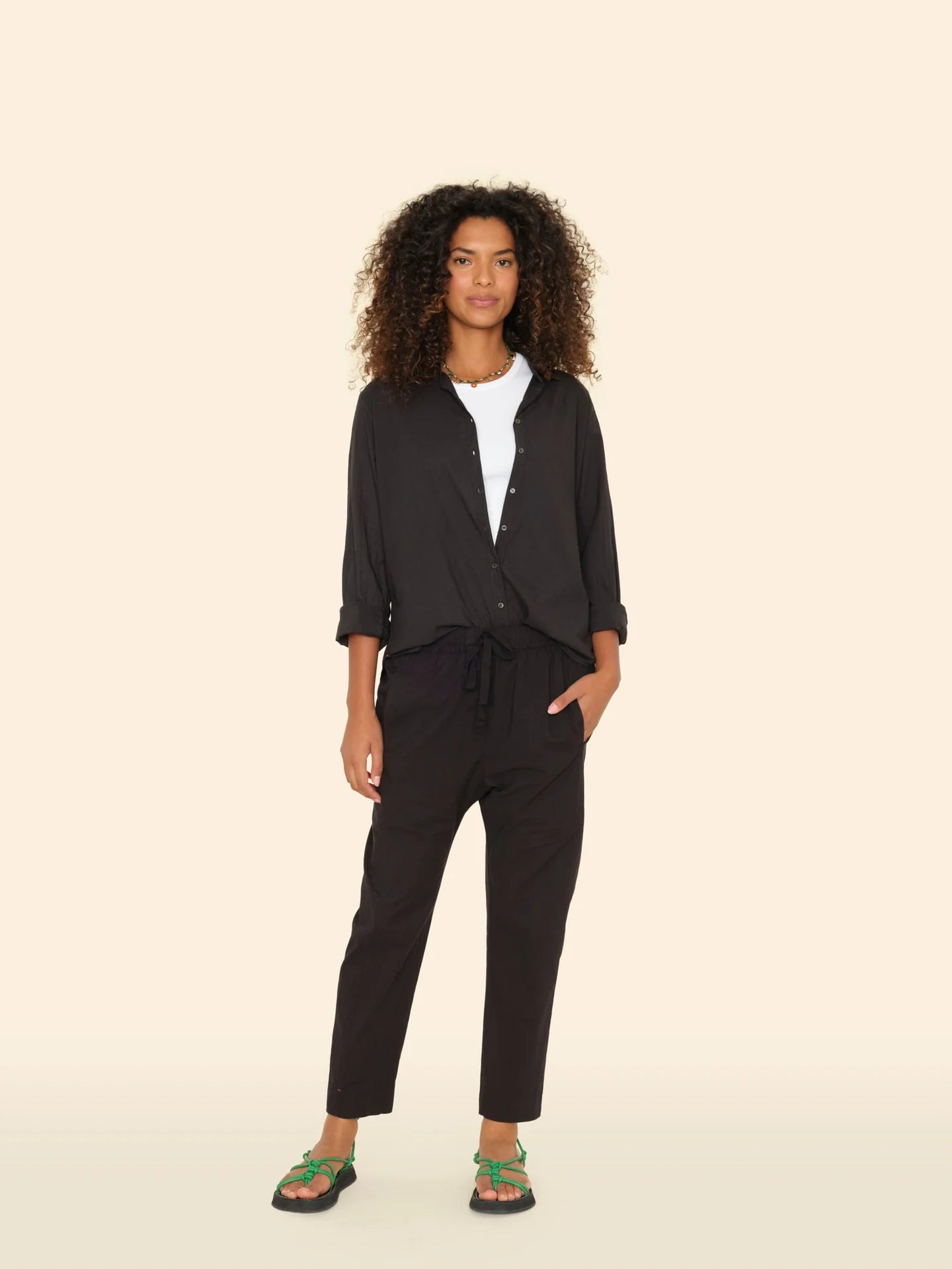 A woman with curly hair wears the Xirena Draper Pant in black, paired with a black lightweight cotton chambray button-up over a white top and black sandals with green straps, standing against a plain beige background.