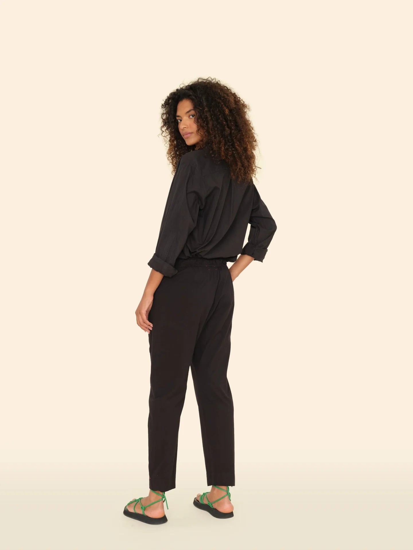 A woman with curly hair stands sideways and looks over her shoulder, wearing a black long-sleeve shirt, Xirena&#39;s Draper Pant in black with side seam pockets, and green sandals—all set against a plain beige background.