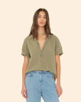 A woman with long hair is seen in an olive green Channing Shirt from Xirena, crafted from lightweight woven cotton, and matched with light blue jeans against a plain beige backdrop. Her look is completed with a colorful beaded necklace, capturing the relaxed elegance typical of Los Angeles design.