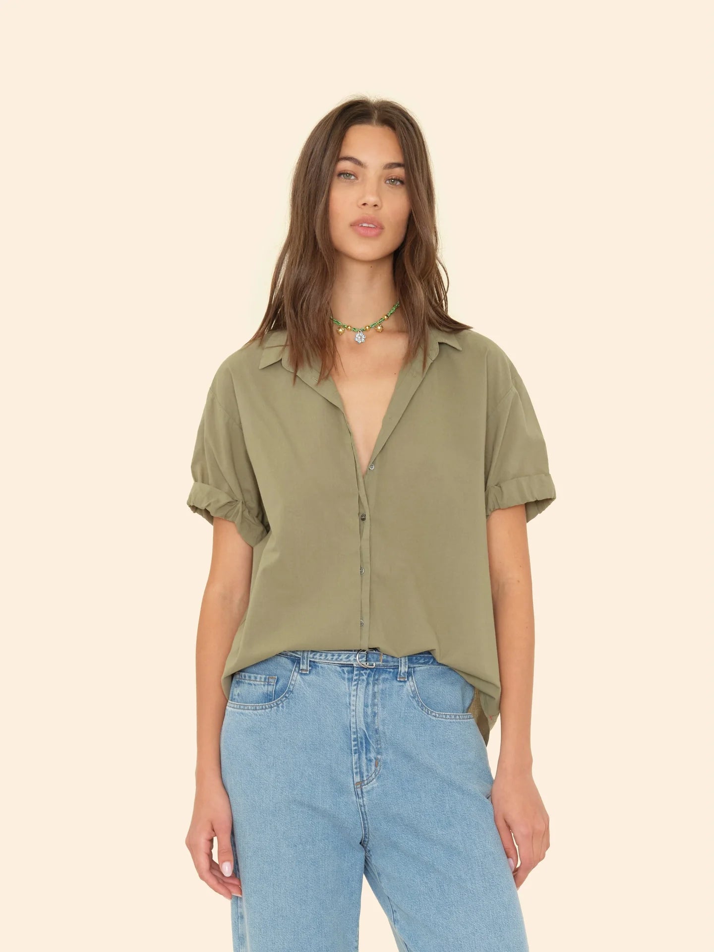 A woman wears the Xirena Channing Shirt S25—an olive green, short-sleeve cotton chambray button-up—paired with high-waisted light blue jeans and a beaded necklace, creating a "boy meets girl" vibe against a beige background.