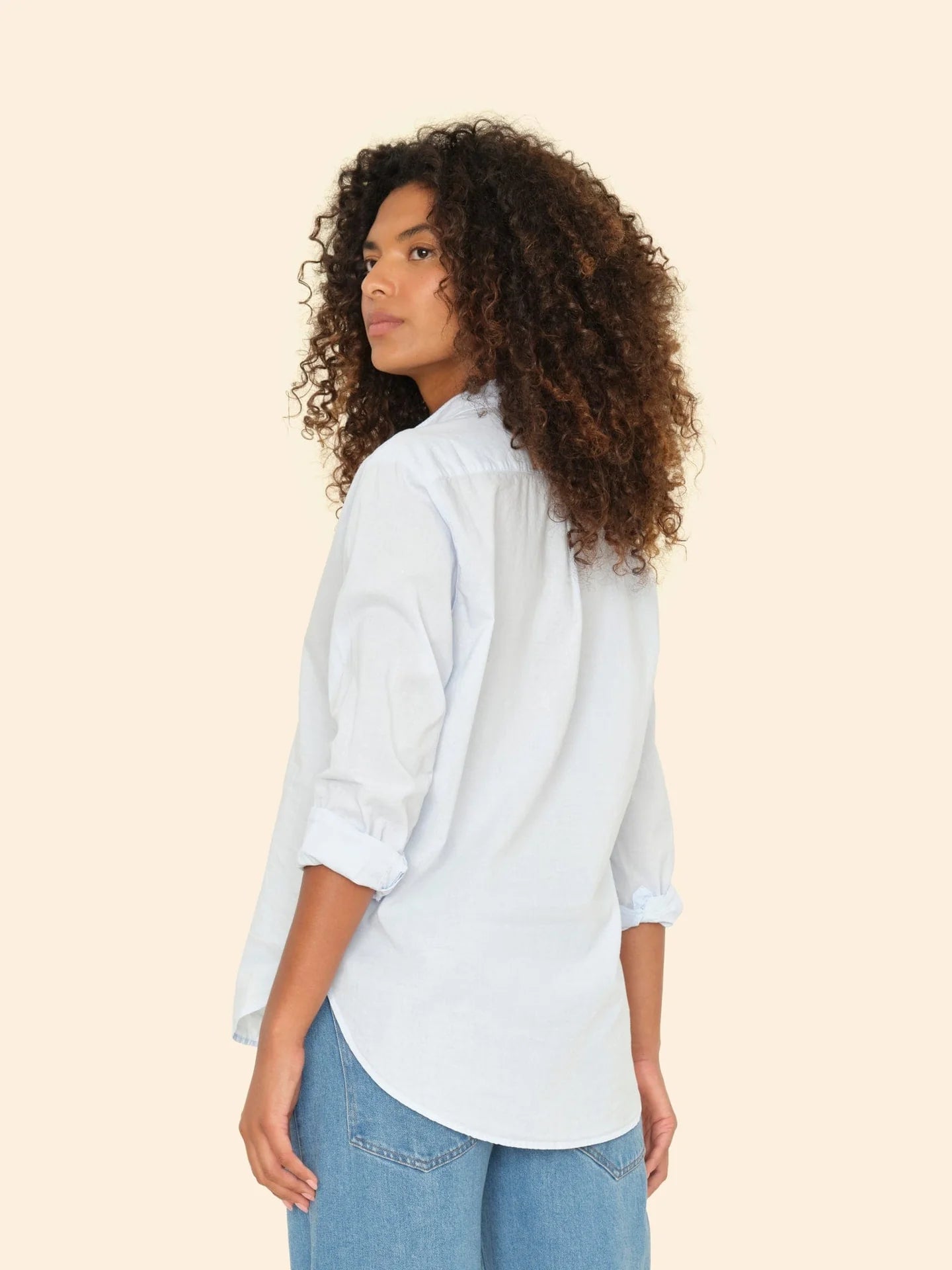 A person with curly hair is seen from behind, wearing a light blue Beau Shirt by Xirena, featuring rolled-up sleeves and classic blue jeans. This menswear-inspired look is crafted from soft cotton poplin, set against a plain, light beige background.