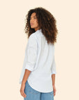 A person with curly hair is seen from behind, wearing a light blue Beau Shirt by Xirena, featuring rolled-up sleeves and classic blue jeans. This menswear-inspired look is crafted from soft cotton poplin, set against a plain, light beige background.