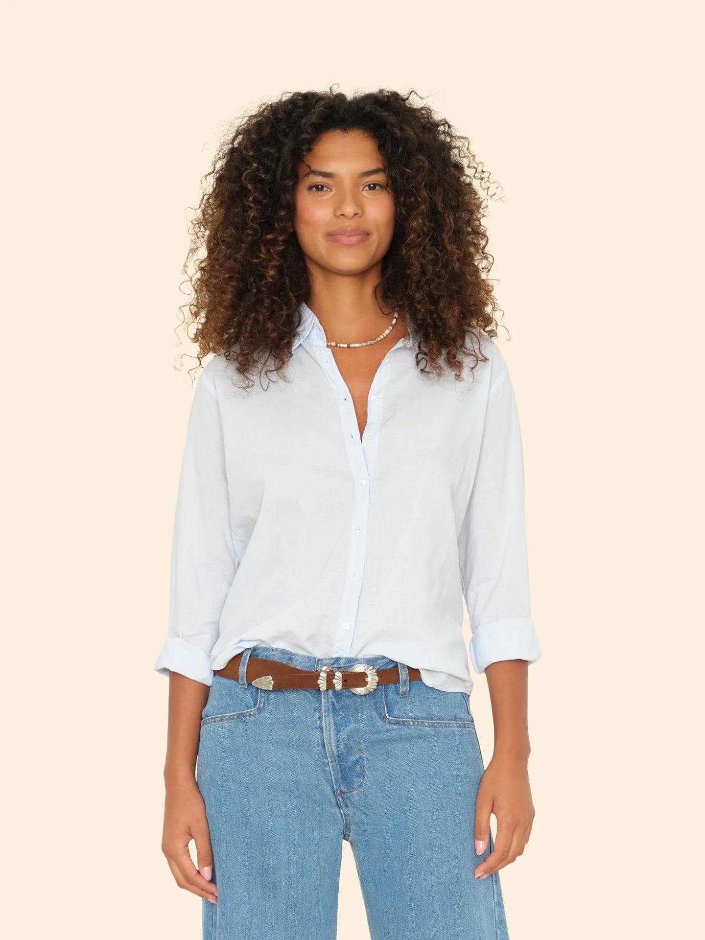 A person with curly hair is wearing the Beau Shirt by Xirena, made from cotton poplin, with the sleeves casually rolled up. Paired with blue jeans, they stand against a plain beige background, smiling softly at the camera.