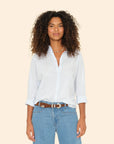 A person with curly hair is wearing the Beau Shirt by Xirena, made from cotton poplin, with the sleeves casually rolled up. Paired with blue jeans, they stand against a plain beige background, smiling softly at the camera.