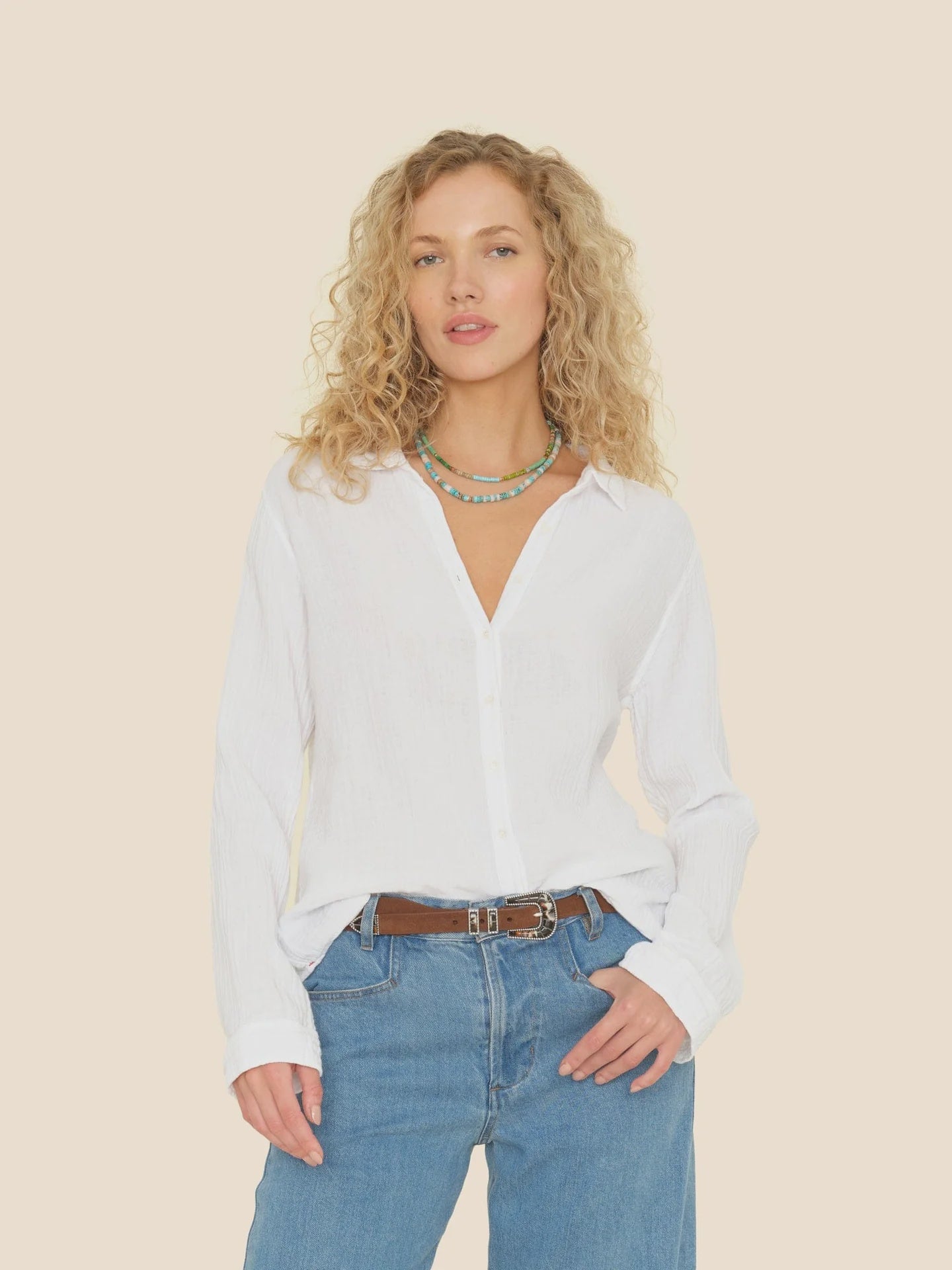 A person with curly blonde hair wears a lightweight Scout Shirt by Xirena in white and blue jeans, standing against a plain background. Accentuated by a colorful beaded necklace and a brown belt, they gaze directly at the camera.