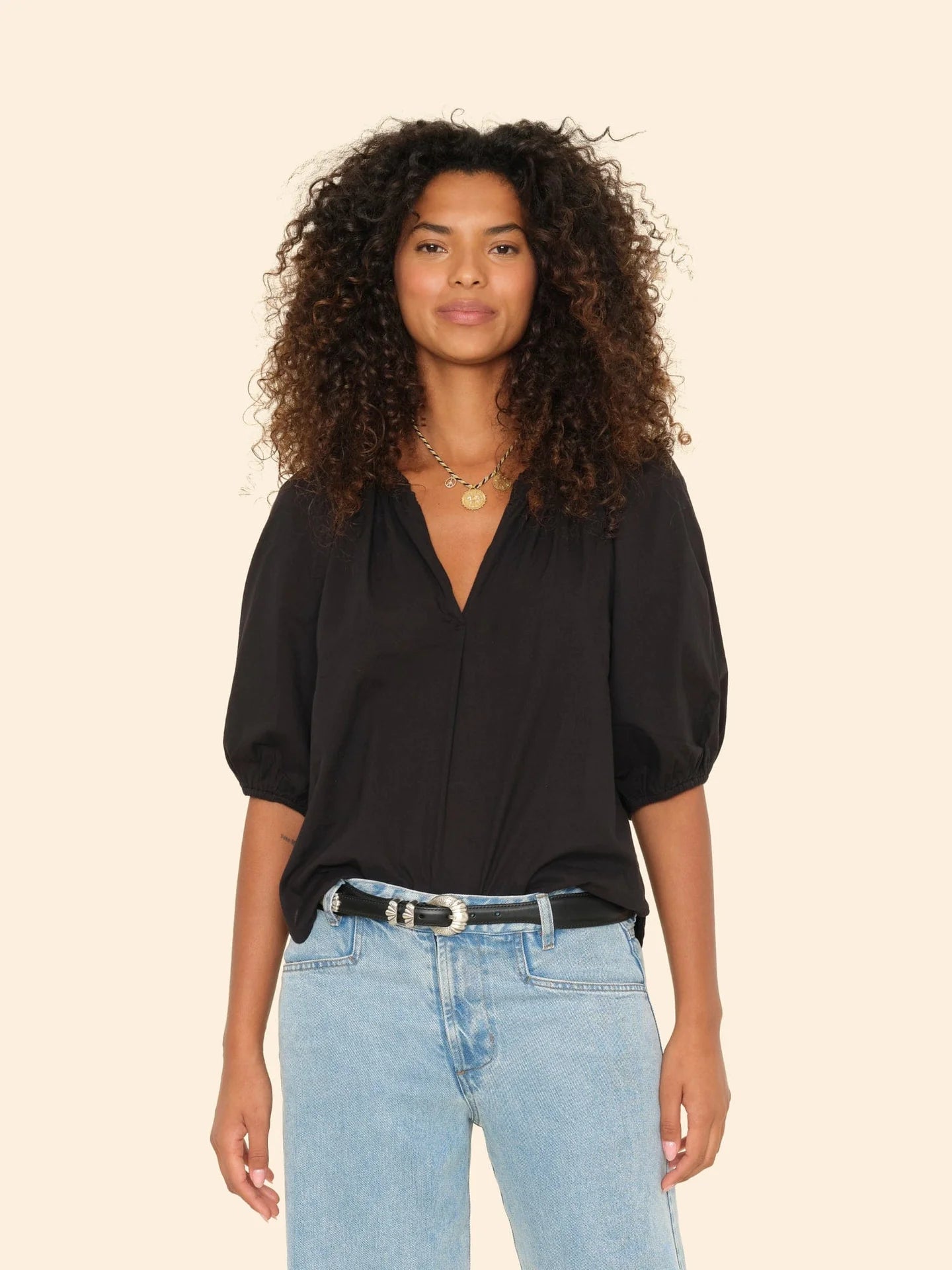 A woman with curly hair wears the black Jules Top with bubble sleeves by Xirena and light blue jeans, accessorized with layered gold necklaces and a black belt, standing against a plain beige background.