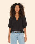 A woman with curly hair wears the black Jules Top with bubble sleeves by Xirena and light blue jeans, accessorized with layered gold necklaces and a black belt, standing against a plain beige background.