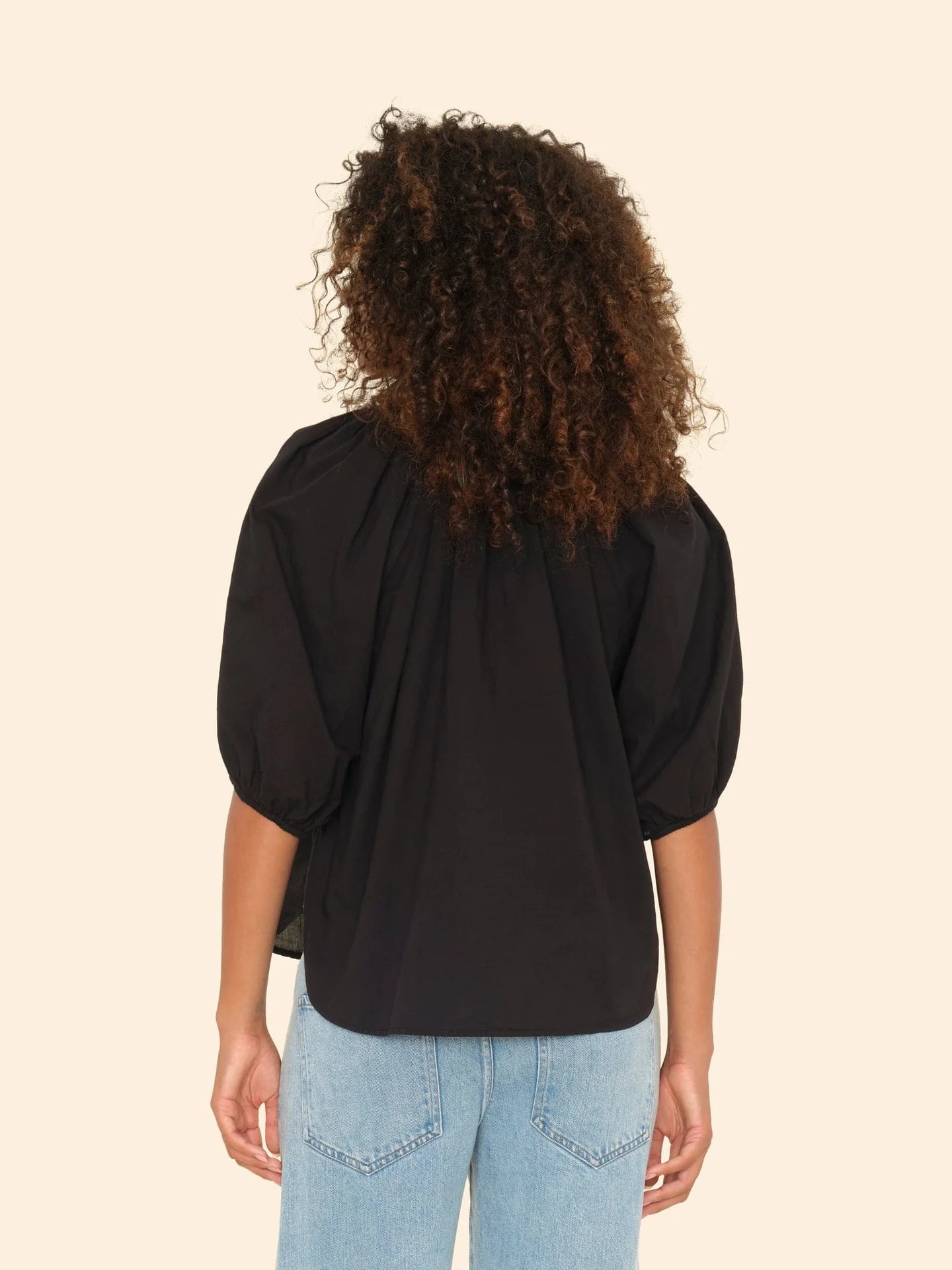 A person with curly hair is seen from the back wearing the Xirena Jules Top, a black cotton poplin blouse with bubble sleeves, and light blue jeans against a beige background.