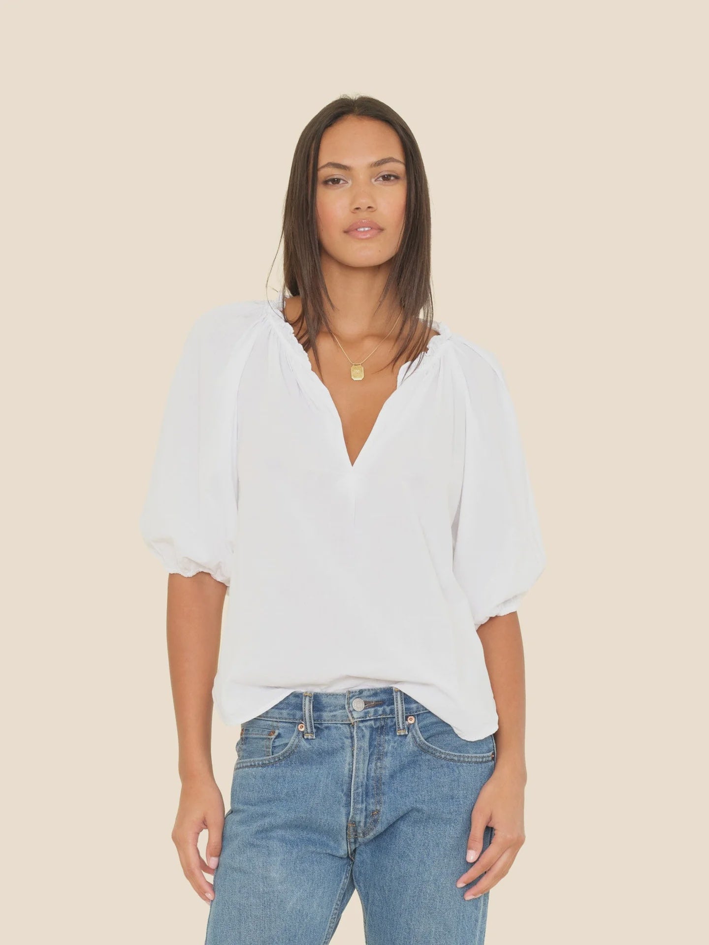 A woman with straight, shoulder-length hair and a gold necklace wears the Xirena Jules Top—a loose-fitting white cotton poplin blouse with bubble sleeves—paired with blue jeans, standing against a plain beige background.