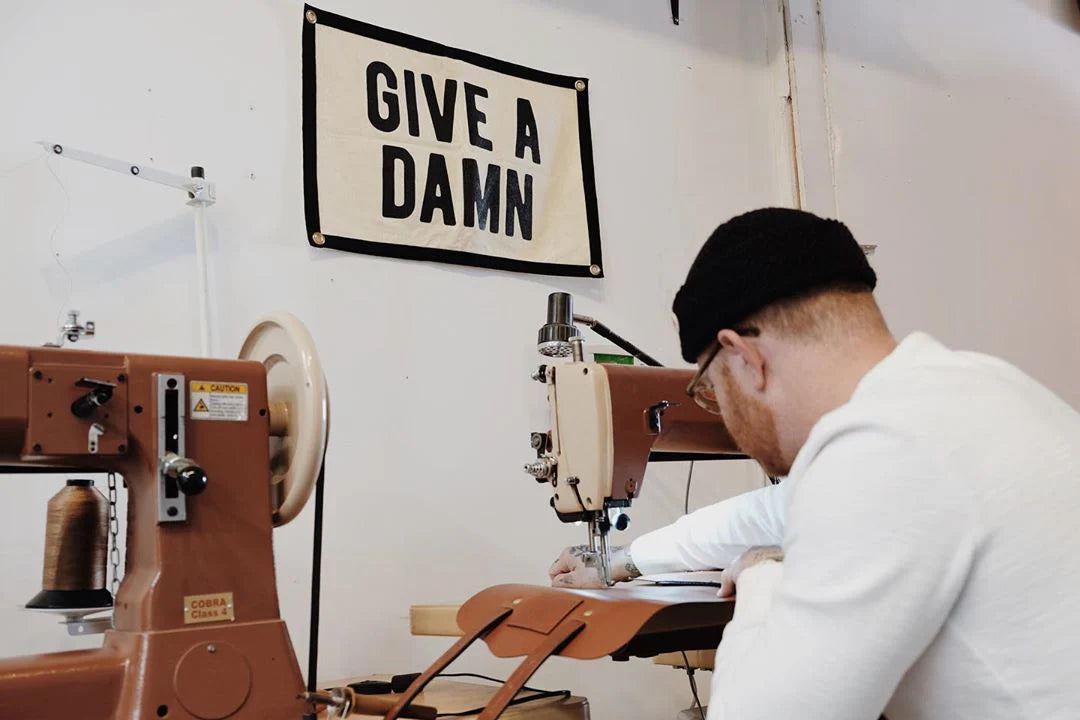 A person in a black beanie works at a sewing machine in a workshop adorned with the Oxford Pennant "Give A Damn Camp Flag 15x22" on the wall. Industrial sewing machines and tools fill the creative space.