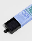 A box labeled "Coastal Forest" by Faire contains neatly arranged black sandalwood Low Smoke Incense sticks, partially pulled out with a black ribbon. The packaging highlights natural ingredients and describes the incense as gentle and refined.