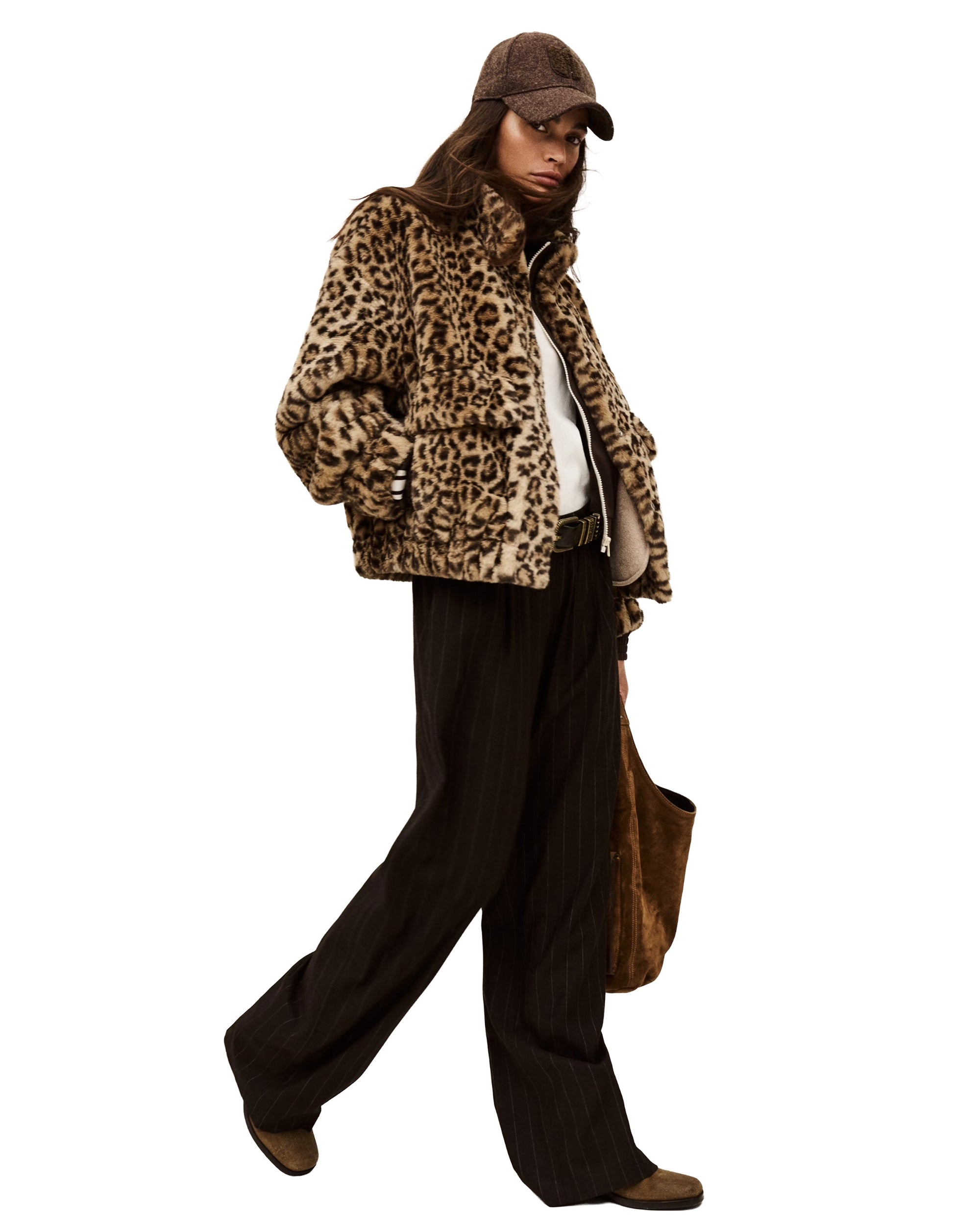 A person stands confidently against a white background, wearing the BA&SH Coat Jaros Leopard, dark wide-leg pants, brown shoes, a baseball cap, and carrying a brown tote bag.