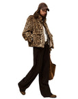 A person stands confidently against a white background, wearing the BA&SH Coat Jaros Leopard, dark wide-leg pants, brown shoes, a baseball cap, and carrying a brown tote bag.