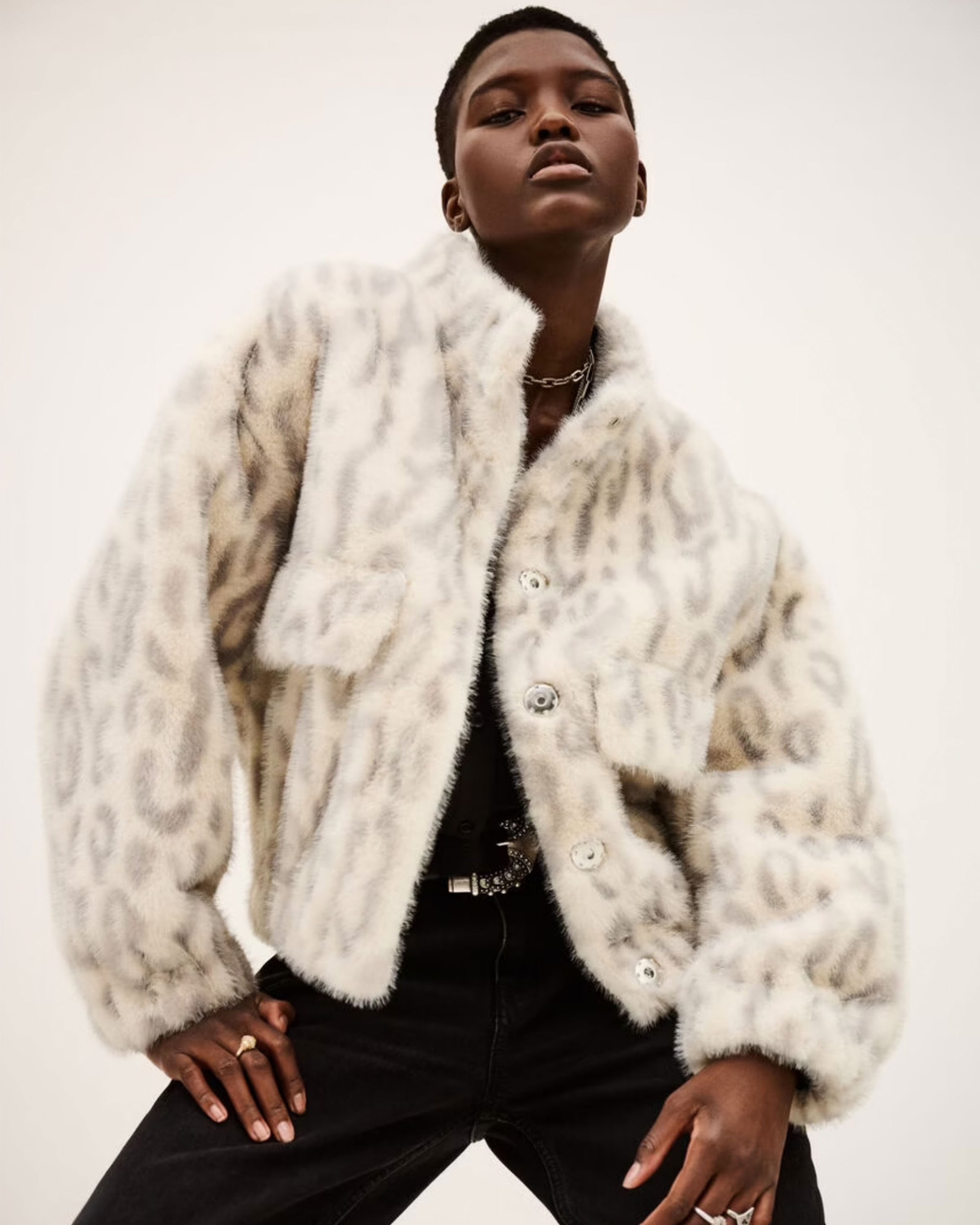 A person poses confidently in the BA&SH Coat Saros, a loose fit, light-colored cropped faux fur jacket with bold animal print and large buttons, paired with black pants against a plain background.