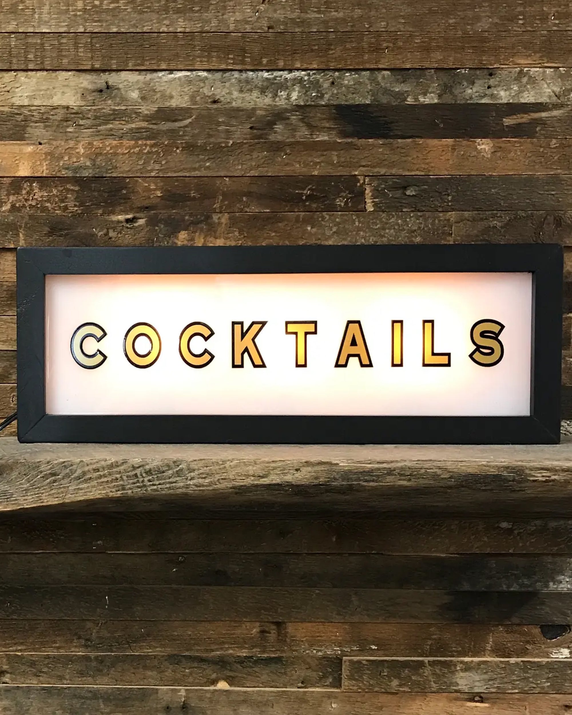 The Faire Lighted Sign is a rectangular, wall-mountable sign displaying "COCKTAILS" in bold black letters, illuminated against a wooden plank wall background.